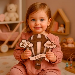 Enfant assise dans sa chambre de bébé avec jouets, tenant une pancarte montgolfière personnalisée