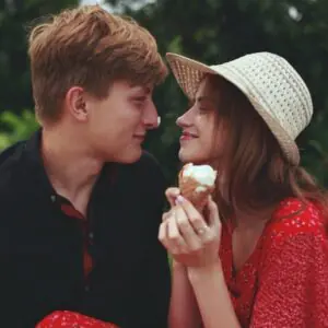 Jeune couple complice en extérieur, elle en robe rouge et chapeau de paille tenant une glace, lui penché vers elle