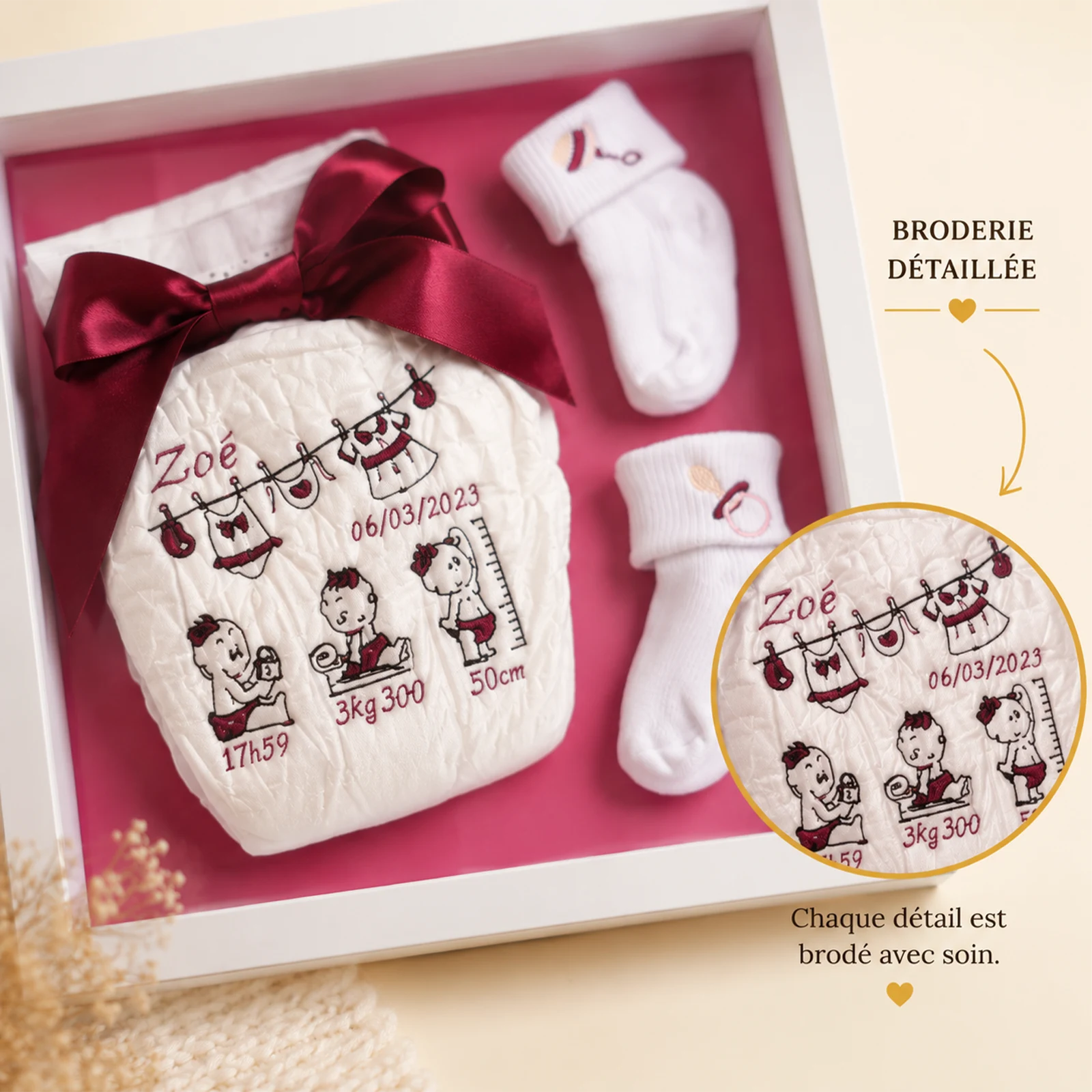 Gros plan de la couche brodée au prénom Zoé, date et mesures de naissance en fil bordeaux, chaussons blancs sur fond rose