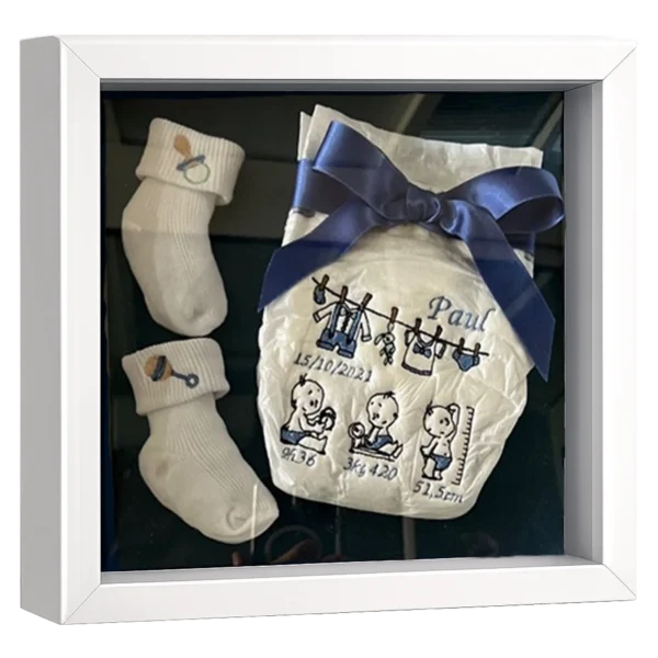 Cadre naissance profond blanc avec chaussons de bébé crème et couche brodée de motifs bleus, prénom et date à personnaliser
