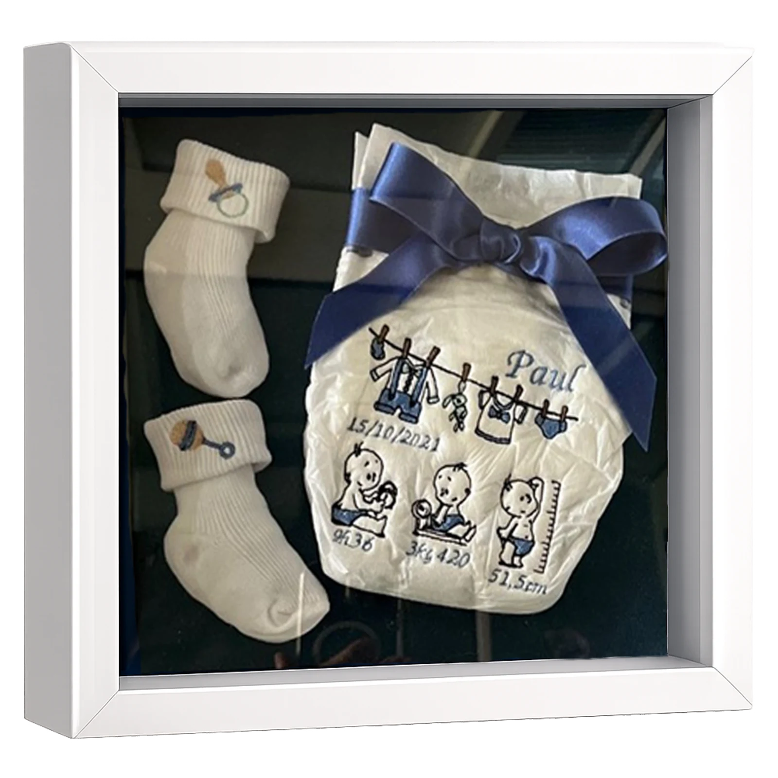 Cadre naissance profond blanc avec chaussons de bébé crème et couche brodée de motifs bleus, prénom et date à personnaliser